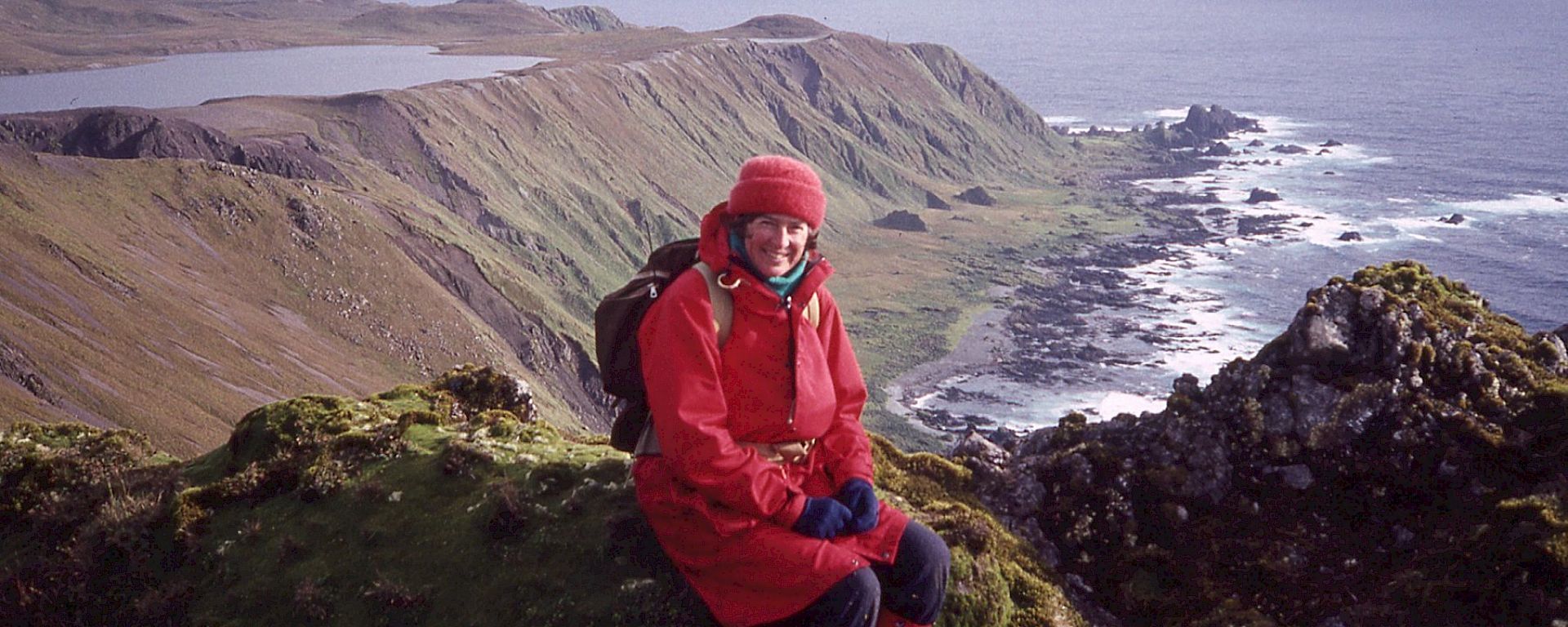 A woman sits on some green mossy rocks with cliffs, rocks and the ocean behind her. The location is Macquarie Island.