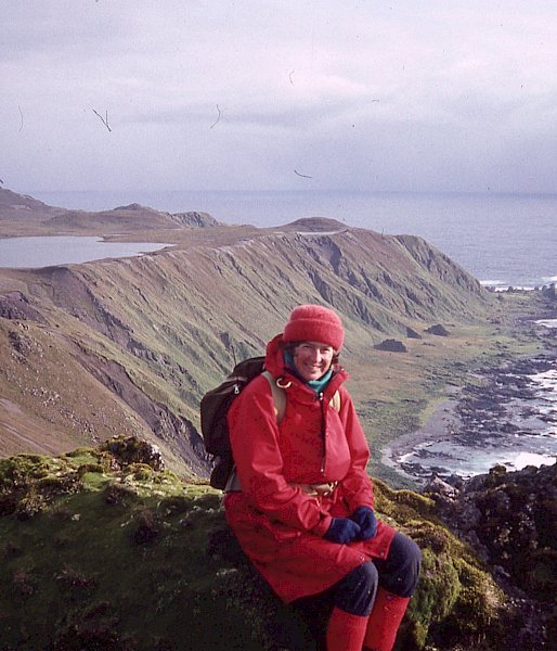 A woman sits on some green mossy rocks with cliffs, rocks and the ocean behind her. The location is Macquarie Island.
