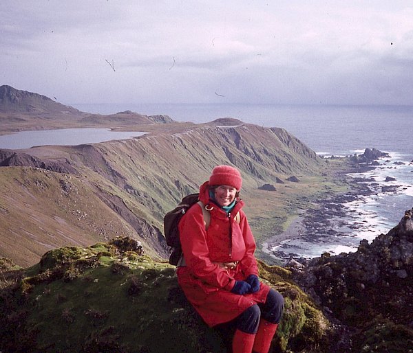 A woman sits on some green mossy rocks with cliffs, rocks and the ocean behind her. The location is Macquarie Island.