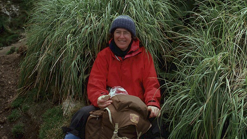 A woman in a red jacket sits in the grass eating lunch.