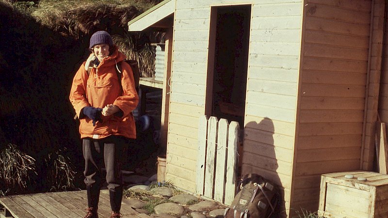 A woman in hiking and survival clothing stands out the front of a wooden field hut and smiles.