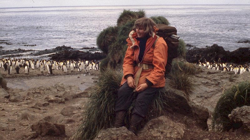 A woman in an orange jacket leans against s grassy rock. Behind her are dozens of king penguins and the ocean