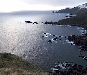 A view down a hill towards the grey ocean .  Waves break over the rocky shore with snow capped mountains above.