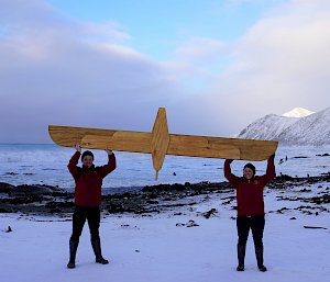 Two people on a snowy beach hold up a wooden albatross