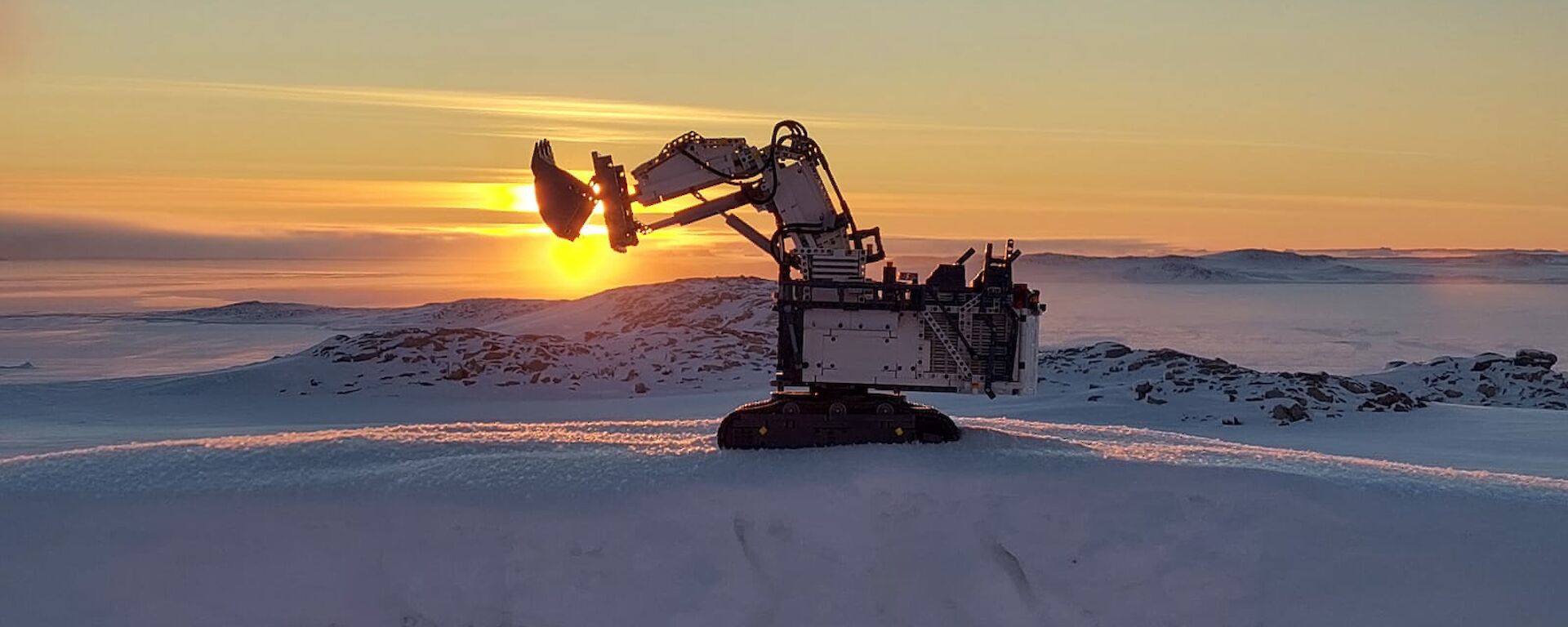 A large earth-digging vehicle made from Lego with its shovel raised parked on snowy ground. There are low hills and sea ice in the background