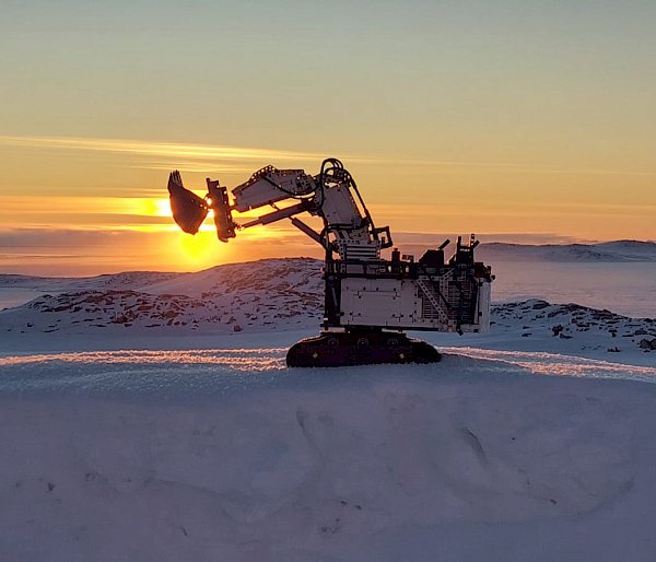 A large earth-digging vehicle made from Lego with its shovel raised parked on snowy ground. There are low hills and sea ice in the background