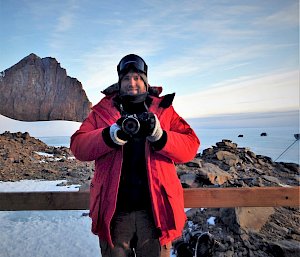 Expeditioner in red goose down jacket stands on hut balcony holding camera with peak of Rumdoodle behind