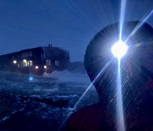 Silhouette of man with head torch on, in background the red shed with lights on