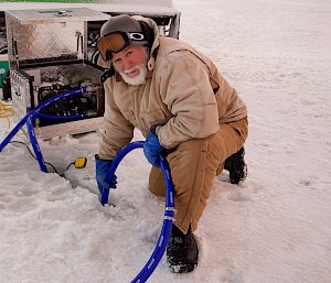 A man kneeling on the ice with a sea ice drill pushed in to a slot in the ice