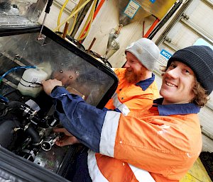 2 plumbers wearing hi-vis working on a drill in a workshop