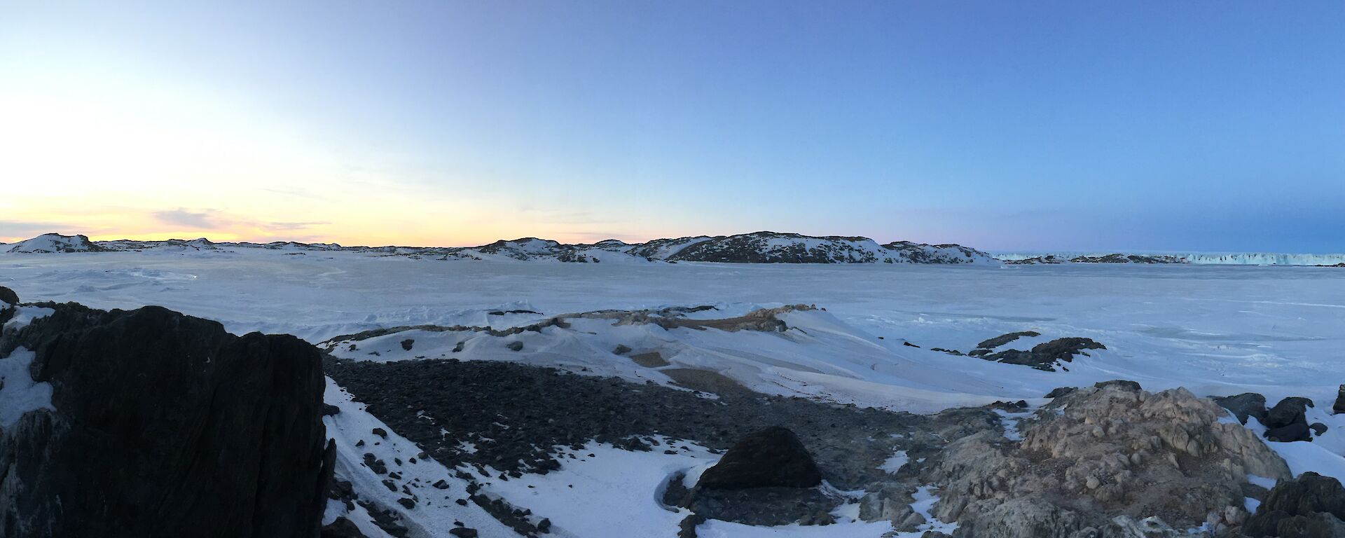 Panoramic view of ice and snow covered hills with a blue sky and the sun just below the horizon
