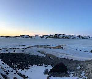 Panoramic view of ice and snow covered hills with a blue sky and the sun just below the horizon