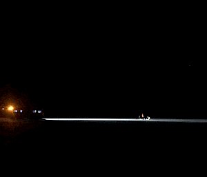 A Hagglund vehicle casts its headlights across a dark landscape highlighting some people just ahead in the darkness