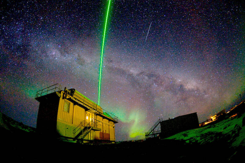 A green laser beam points up into the night sky from inside a small building.