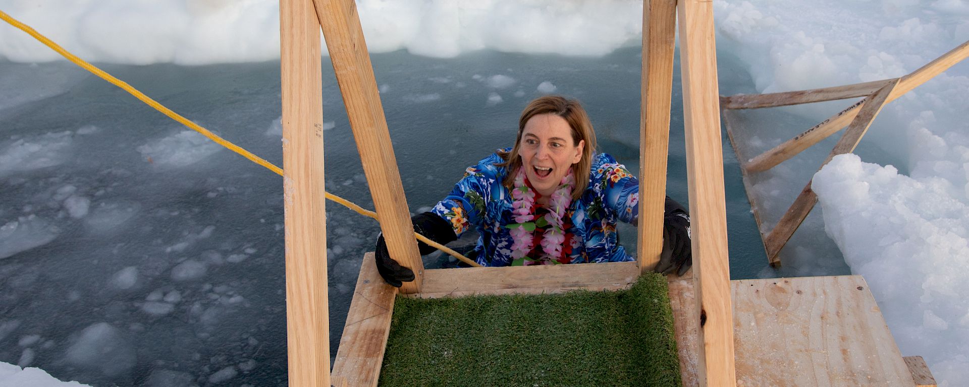 A woman in a Hawaiian shirt climbs out of an ice hole