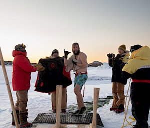 A man smiles at the camera surrounded by a group of expeditioners, with an Antarctic background