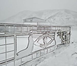 A snow covered fence sits in front of a building and hill