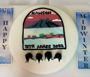 Large cake iced in white icing with Mawson 75th logo on top, beside sign reads 'happy midwinter'