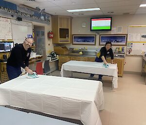 Two men standing ironing tablecloths on two tables side by side