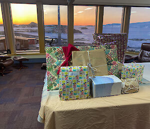 Large pile of gifts on end of pool table, with view of sea-ice and sunset behind through windows