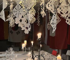 Large dining table set with cutlery, glasses, candles and above paper snowflakes and fairy lights shining