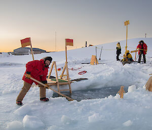 Man on edge of sea-ice hole, skimming the water with a wooden rake to remove sea-ice forming on the surface