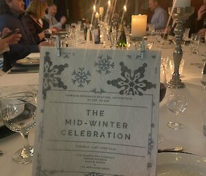 Dinner menu held up to camera, in the background table set with candles, glasses, cutlery and people sitting at their places