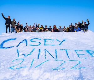A view from below of a group of about 30 people, some with their arms raised, standing on the flat top of a pyramid several metres high and sculpted from snow. On the front face of the pyramid, the words "CASEY WINTER 21/22" are painted in blue snow