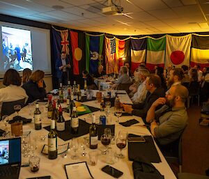 A group of about 30 people in neat attire, seated around 2 dining tables in a room hung with international flags. They are listening to a speech being given by a man standing at the front of the room beside a projector screen