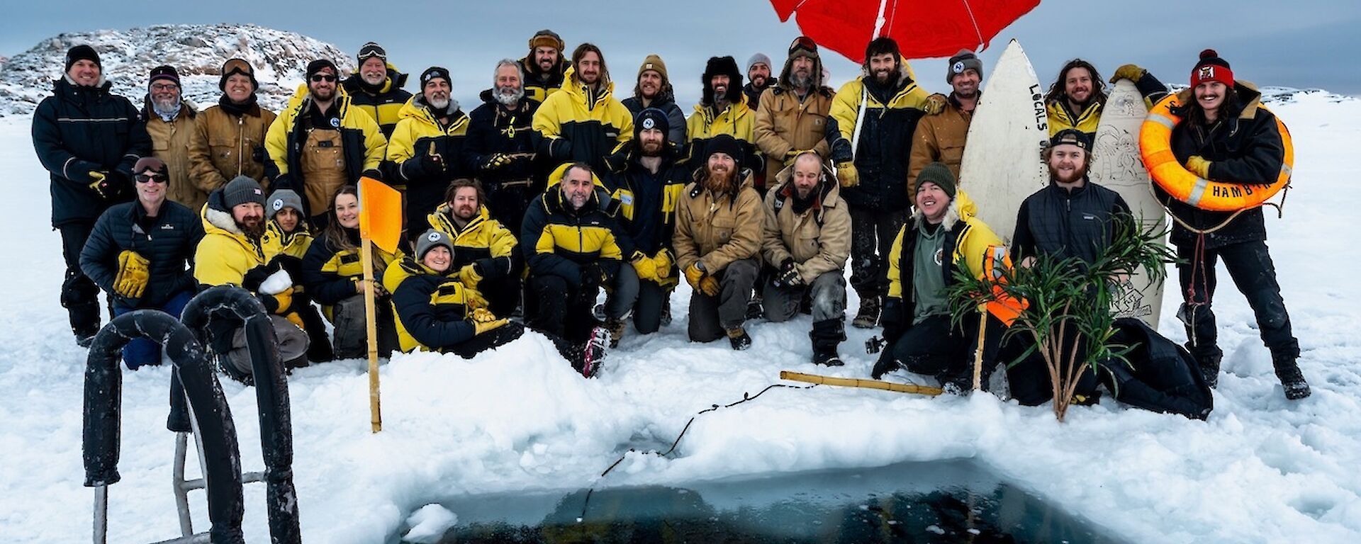 A group of about 30 people in Antarctic weatherproof clothing, arranged in 2 rows sitting, kneeling and standing by a square-shaped pool of water in a wide expanse of sea ice. A red beach and umbrella and a couple of surfboards are set up near the pool's edge for decoration