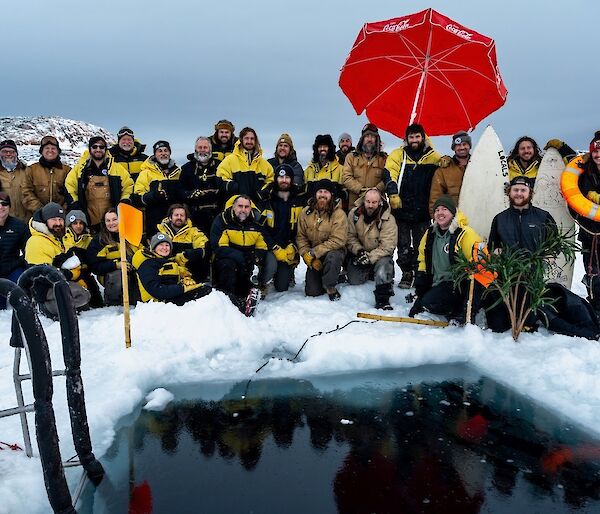 A group of about 30 people in Antarctic weatherproof clothing, arranged in 2 rows sitting, kneeling and standing by a square-shaped pool of water in a wide expanse of sea ice. A red beach and umbrella and a couple of surfboards are set up near the pool's edge for decoration