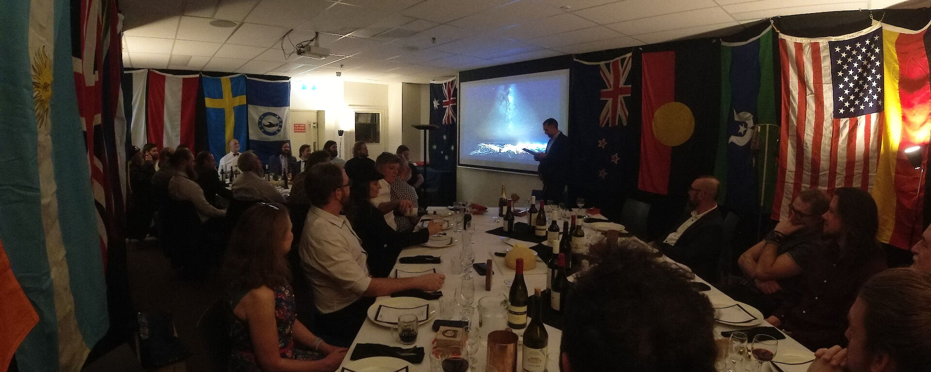 A group of about 30 people in neat attire, seated around 2 dining tables in a room hung with international flags. They are listening to a speech being given by a man standing at the front of the room beside a projector screen