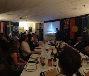 A group of about 30 people in neat attire, seated around 2 dining tables in a room hung with international flags. They are listening to a speech being given by a man standing at the front of the room beside a projector screen