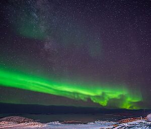 The aurora australis, appearing as a band of bright green light above a bank of low cloud on the horizon. Above the aurora is a starry night sky and the cloudy, patchy haze of the Milky Way