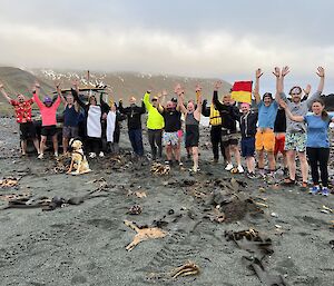 A group of people stand on the grey sandy beach with their hands in the air.