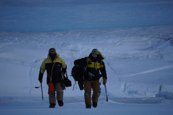 Two men walk towards camera, in the distant background the rolling edge of a glacier meeting the sea ice