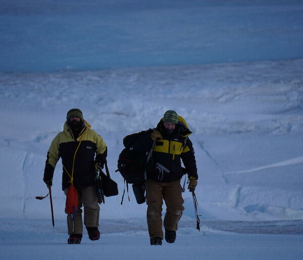 Two men walk towards camera, in the distant background the rolling edge of a glacier meeting the sea ice