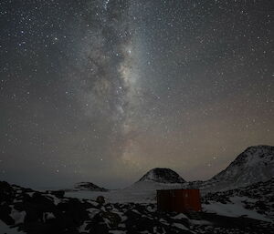 Rocky foreground with hut in shadow, above the expanse of night sky with bright stars and milky way