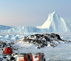 View from elevation on island shows apple hut and two haglunds in foreground and in the distance an overturned iceberg which has a large pointed peak