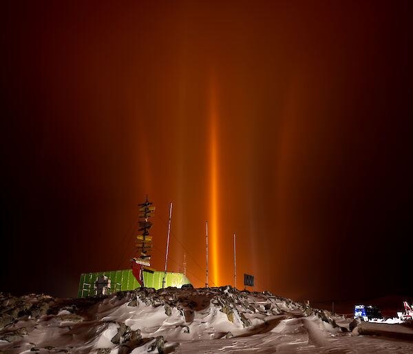 A night scene at an Antarctic station, with a large green shed, a signpost and some parked vehicles in a snowy, rocky area. Some station lights appear as spears of orange light reaching high into the air