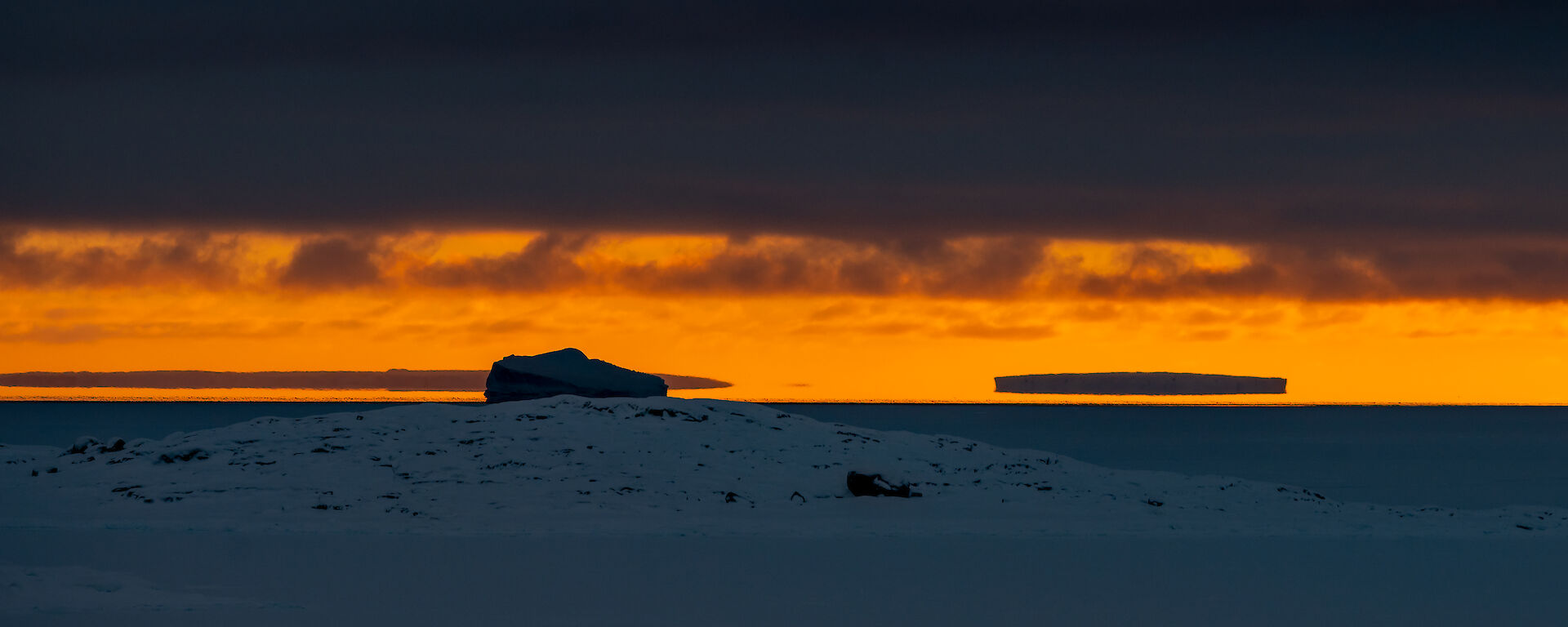 A sunset scene, with bright amber sky showing between a dark layer of cloud and a grey, ice-covered ocean. Icebergs at the horizon appear to be suspended in the sky