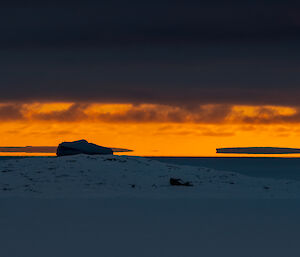 A sunset scene, with bright amber sky showing between a dark layer of cloud and a grey, ice-covered ocean. Icebergs at the horizon appear to be suspended in the sky