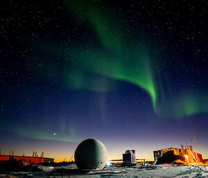 The aurora australis in the night sky above the buildings and structures of an Antarctic station. It looks like diaphanous green ribbons waving above fading violet sunset light