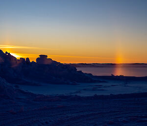 Piles of snow tinted blue in the shadow of the sun setting behind them. A small piece of the sun's halo effect appears like a short beam of light standing beside the snow pile