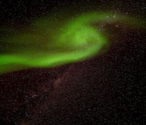 A fragment of the aurora australis in a densely-starred night sky. It looks like a swirl of green light surrounded by a faint red glow against the darkness