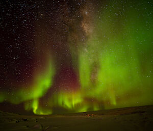 A strong aurora display in the night sky over snowy ground. About two thirds of the sky in this picture is filled with aurora light, appearing like blurred green fire crested with dull red. The dense cloudiness of the Milky Way can be seen behind the aurora