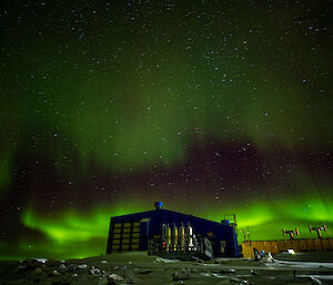 The aurora australis filling the night sky above Antarctic station buildings. Near the horizon the aurora light is in thick skeins of vivid green; just above this is a dark reddish band, which melds into a fainter green, broad swathe of light sweeping upwards