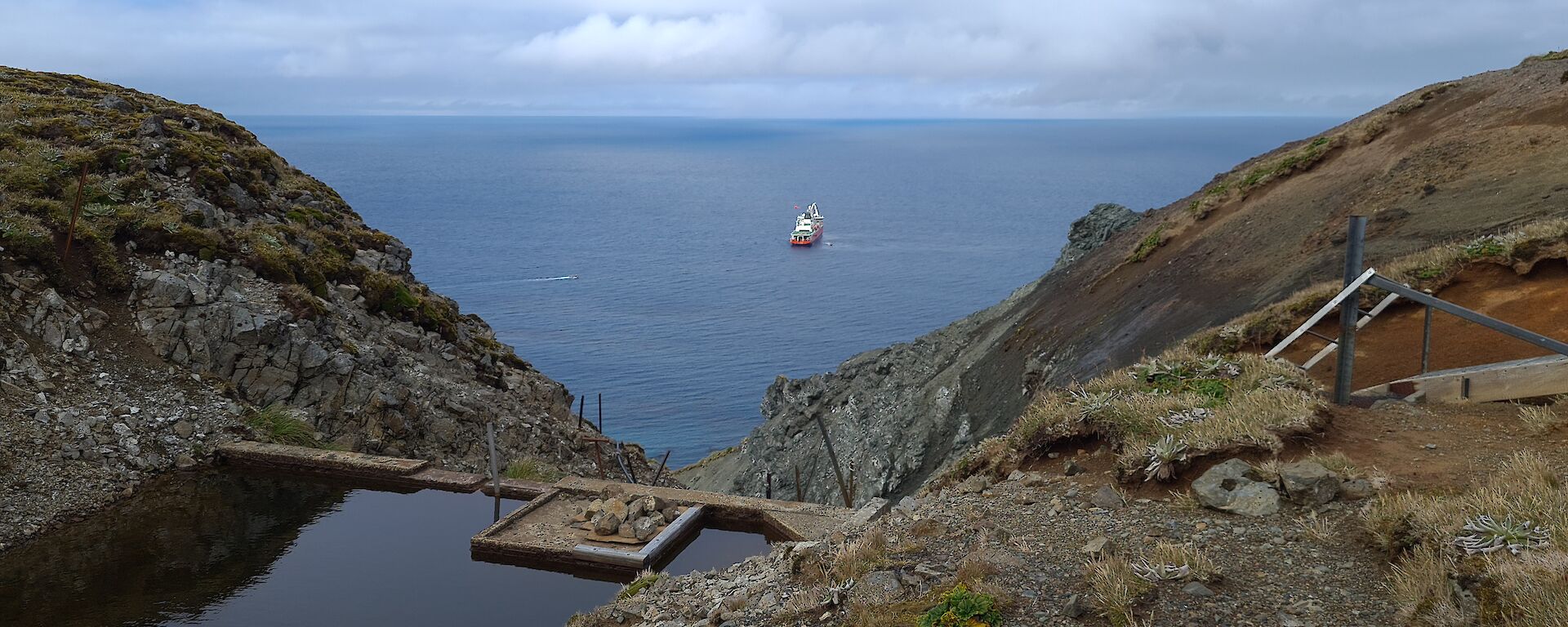 A grassy valley with a water holding dam and the ship Nuyina in the background