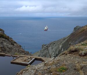 A grassy valley with a water holding dam and the ship Nuyina in the background