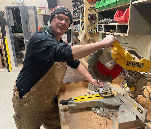 Man in overalls, wearing St Kilda football beanie and safety glasses, holds a band saw and looks to camera and smiles